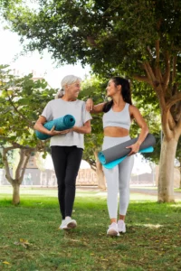 uas mulheres caminhando juntas em um parque enquanto carregam tapetes de yoga enrolados. Uma delas é mais velha, com cabelos grisalhos presos, vestindo uma camiseta cinza e calça preta. A outra é mais jovem, com cabelos escuros presos em um rabo de cavalo, vestindo um conjunto esportivo claro. 